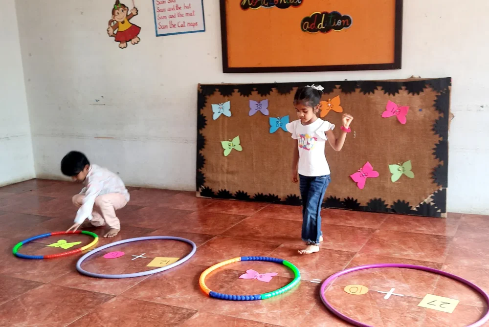 Concentrated UKG children solving horizontal addition problems with paper shapes on the floor, demonstrating innovative learning at Vidhya Peetham i-School.
