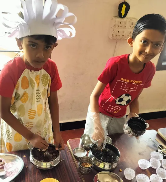 Two young students in chef attire measuring ingredients for chocolate cake making at an i-School activity.