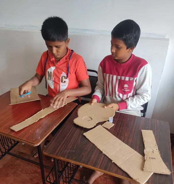 Two students cutting cardboard shapes for a guitar in the Making Musical Instruments activity at Vidhya Peetham i-School.