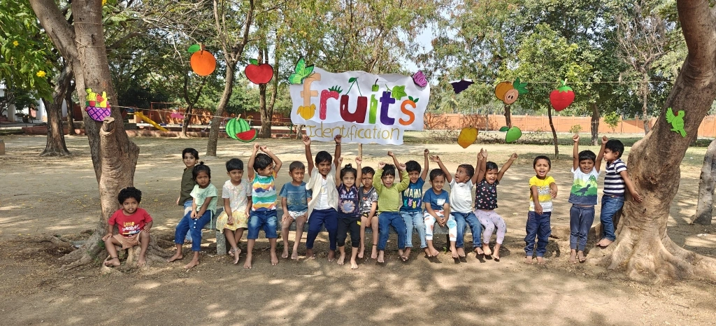 Enthusiastic pre-KG students at Vidhya Peetham i-School sitting under trees decorated with colorful fruit identification cards during fruit identification activities
