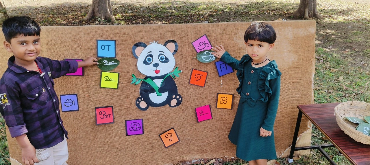 Two students engaging with colorful Tamil letter cards around panda mascot display board