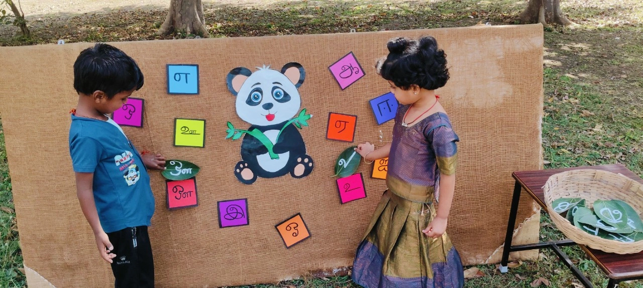 Student selecting Tamil letters from a basket while learning உயிர் எழுத்துகள் at outdoor activity station